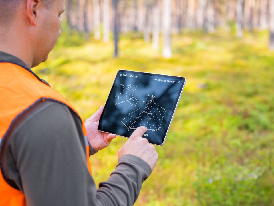 Työvaatteisiin pukeutunut mies katsoo metsässä tablettia.