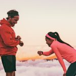 A man and a woman dressed in sportswear are standing in front of a mountainous landscape, looking at a smartwatch on their wrists.