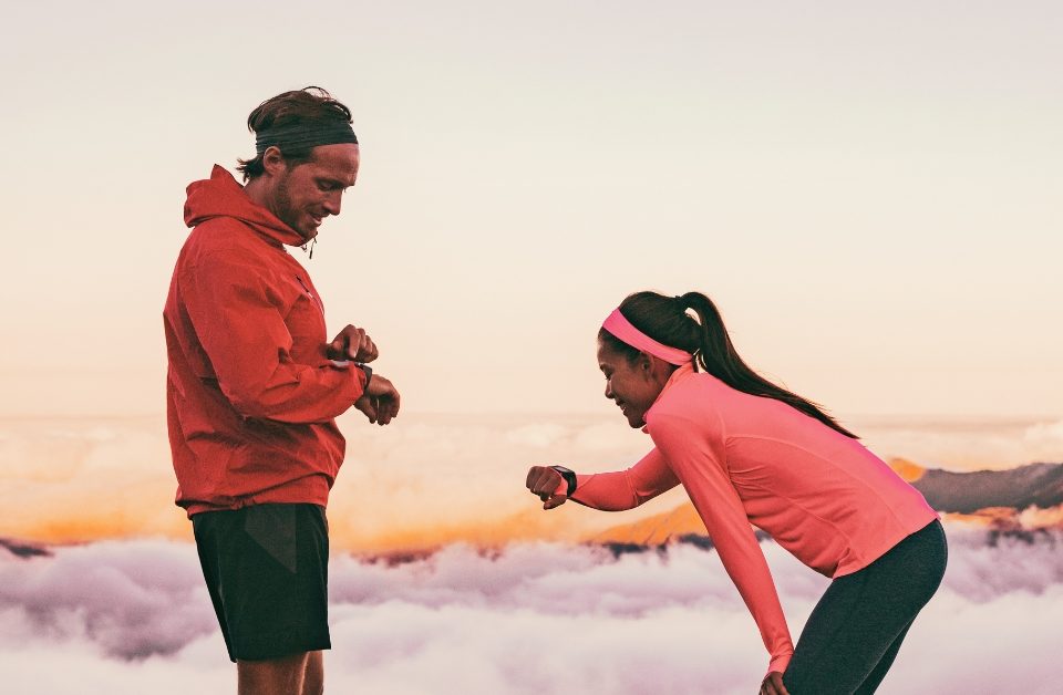 A man and a woman dressed in sportswear are standing in front of a mountainous landscape, looking at a smartwatch on their wrists.