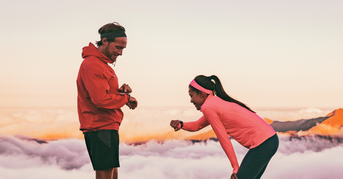 A man and a woman dressed in sportswear are standing in front of a mountainous landscape, looking at a smartwatch on their wrists.