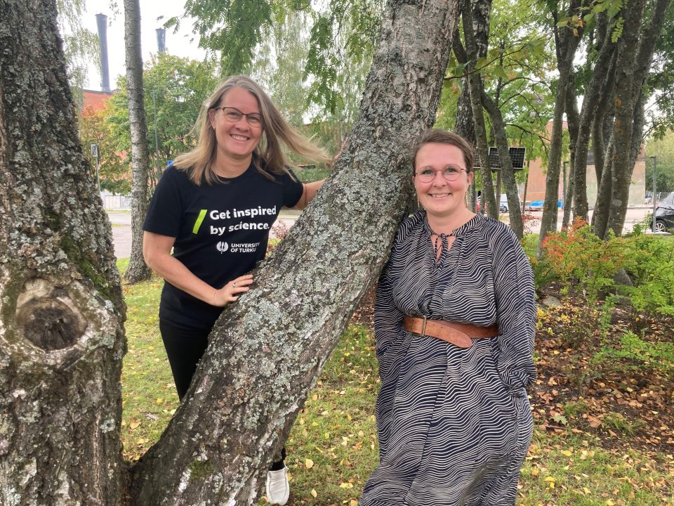 Niina Käyhkö ja Sanna Jokela nojaavat puunrunkoon puistomaisessa maisemassa.