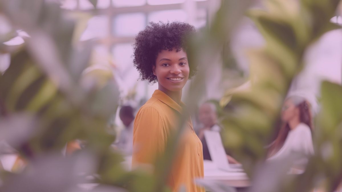 A smiling woman stands in an open-plan office, surrounded by green indoor plants.