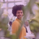 A smiling woman stands in an open-plan office, surrounded by green indoor plants.