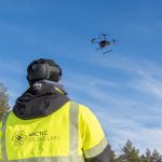 A person flying a drone wearing a jacket that reads Arctic Drone Labs.