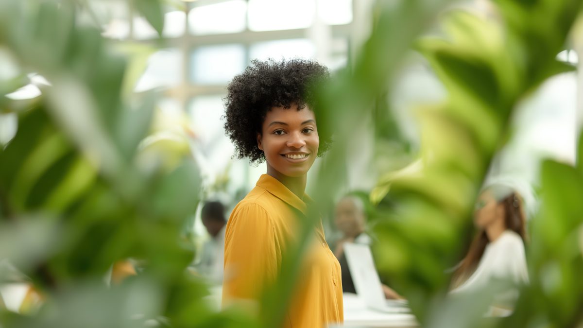 A young woman standing in an open office space, smiling and holding her phone while surrounded by team members working on their laptops at white desks with light gray accents. The scene is captured from the perspective of someone sitting down behind one desk looking towards them. She's wearing casual attire, such as pants or jeans paired with an orange blouse. Green plants add a modern feel to it all.