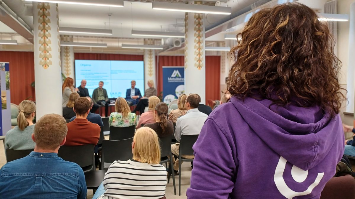 Kuva yleisöstä Lajitiedon käyttö ja tulevaisuus -seminaarista. Etualalla henkilö violetissa hupparissa ja taustalla paneelikeskustelu.
