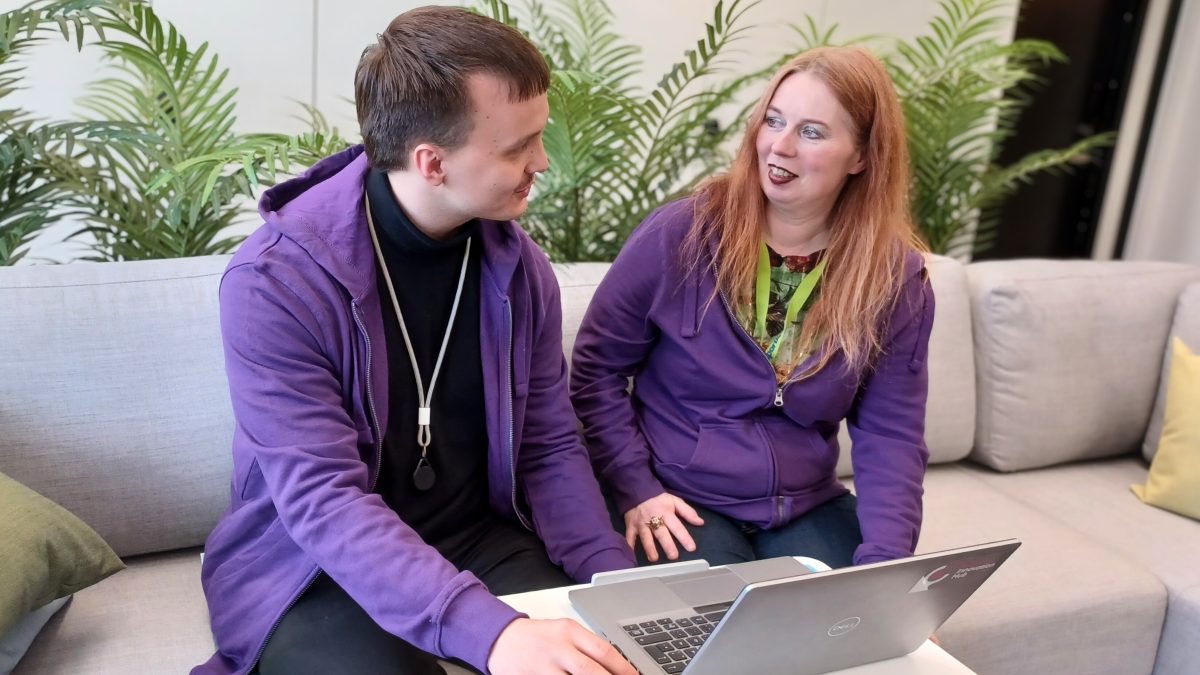Two people wearing purple hoodies are sitting on a light grey sofa, looking at a laptop on a small white table. There are green plants in the background.