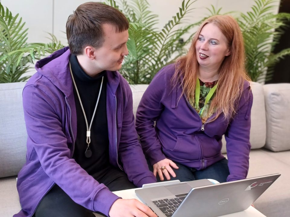 Two people wearing purple hoodies are sitting on a light grey sofa, looking at a laptop on a small white table. There are green plants in the background.
