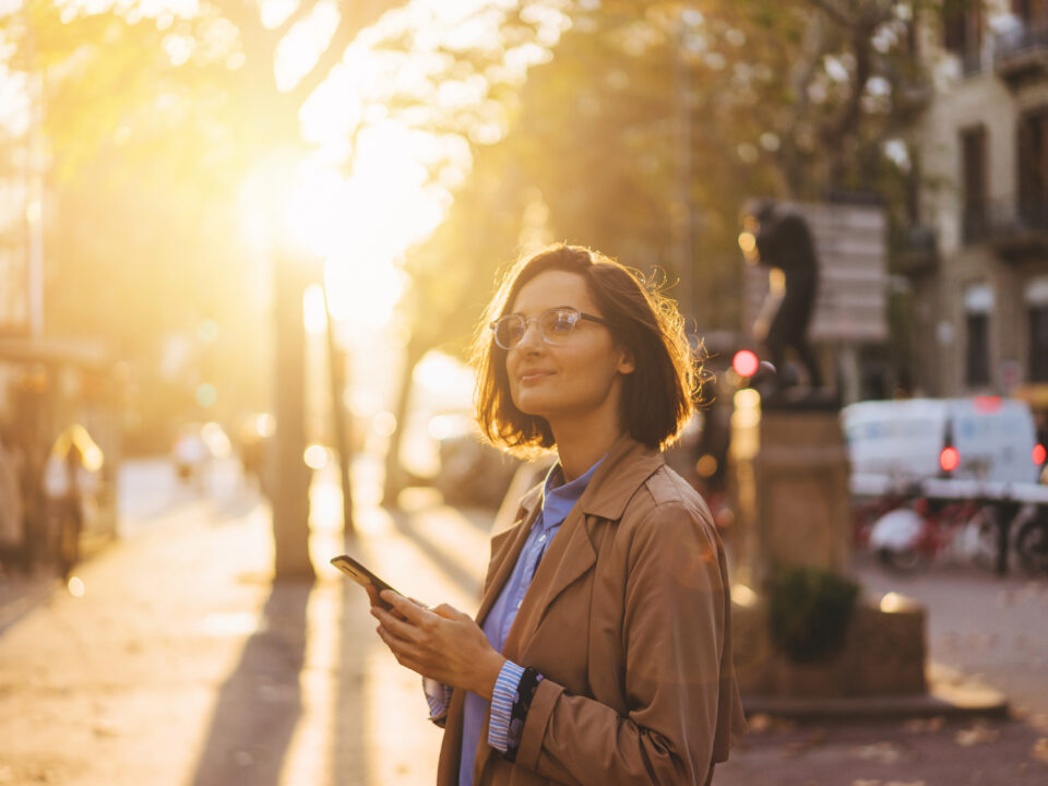 A person standing on a city street at sunset, looking at a smartphone. Trees, buildings, and a statue are visible in the background, with warm golden light illuminating the scene.