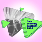 Graphic with a pink background featuring a triangular black‑and‑white photo of an audience looking toward a bright stage. Next to it is a green triangular shape with dark blue text reading 'Data Economy Spotlight 2026' and smaller text 'Datatalouden kiinnostavimmat'. Several smaller green triangular graphic elements surround the main shapes.