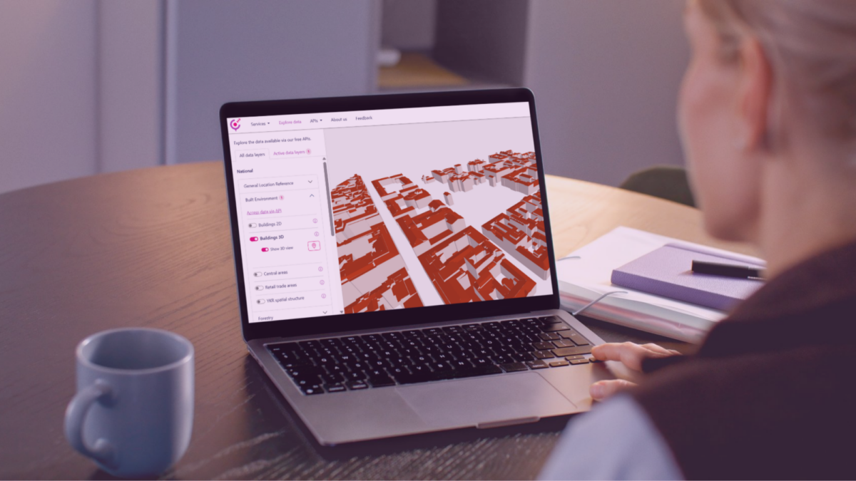 A person working at a desk views a 3D city model on a laptop screen, with notebooks and a coffee mug placed nearby.