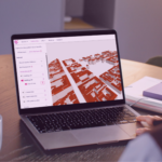 A person working at a desk views a 3D city model on a laptop screen, with notebooks and a coffee mug placed nearby.
