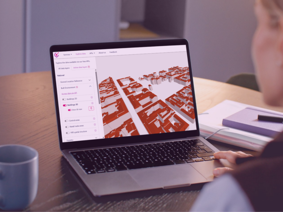 A person working at a desk views a 3D city model on a laptop screen, with notebooks and a coffee mug placed nearby.