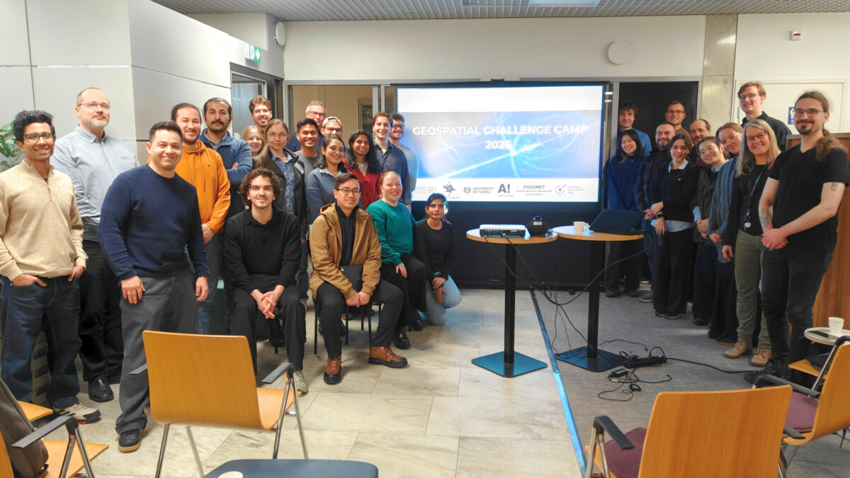 A group of Geospatial Challenge Camp 2026 participants indoors at a midterm event, standing and sitting in front of a presentation screen displaying the text “Geospatial Challenge Camp 2026”.