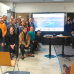 A group of Geospatial Challenge Camp 2026 participants indoors at a midterm event, standing and sitting in front of a presentation screen displaying the text “Geospatial Challenge Camp 2026”.