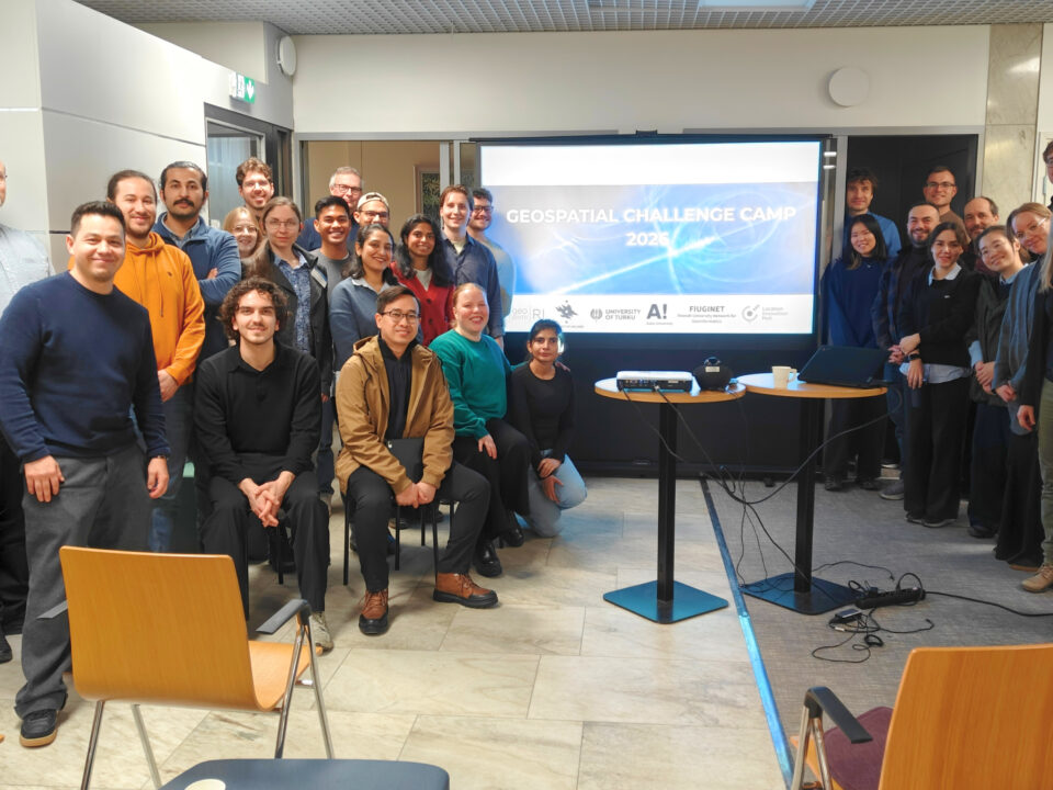 A group of Geospatial Challenge Camp 2026 participants indoors at a midterm event, standing and sitting in front of a presentation screen displaying the text “Geospatial Challenge Camp 2026”.