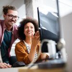 Two people working together at a desk, looking at a desktop computer screen in an open office environment.