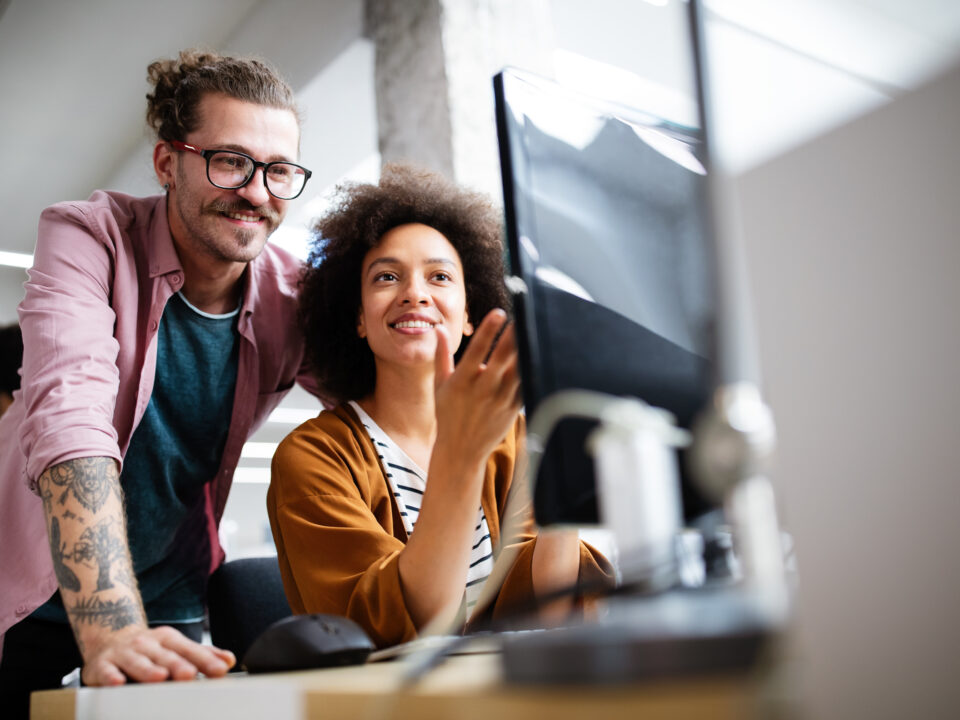 Two people working together at a desk, looking at a desktop computer screen in an open office environment.