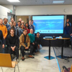 Group photo of participants standing and sitting in a meeting room around a large screen displaying “Geospatial Challenge Camp 2026,” with a projector, laptop, and chairs visible in the foreground.