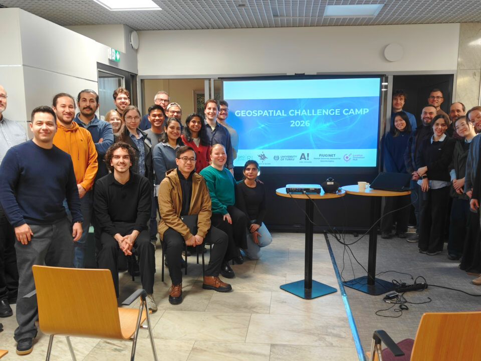 Group photo of participants standing and sitting in a meeting room around a large screen displaying “Geospatial Challenge Camp 2026,” with a projector, laptop, and chairs visible in the foreground.