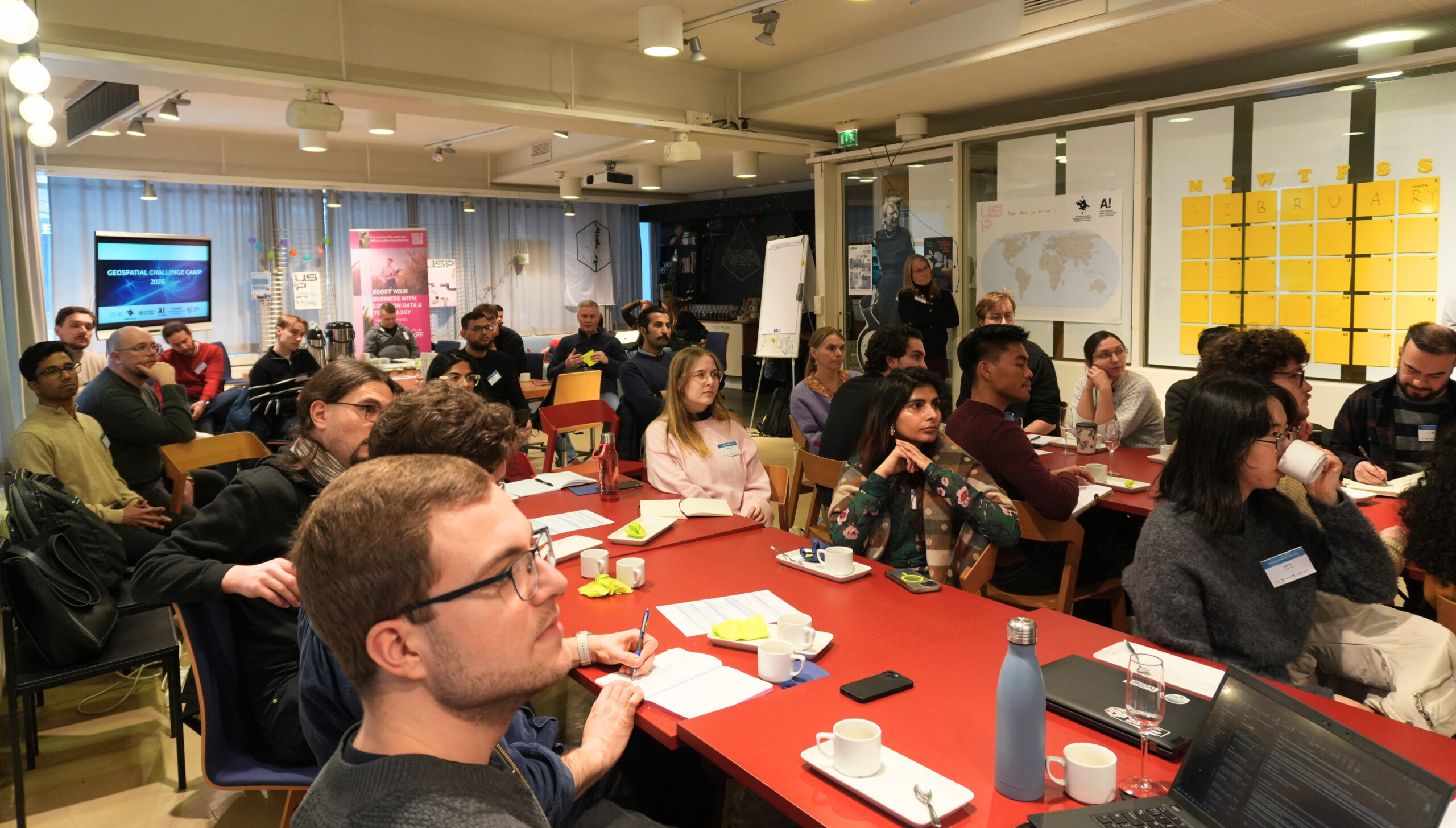 A group of people seated at red tables in a well-lit room, attending a workshop or presentation. A screen displaying “Geospatial Challenge Camp 2026” is visible at the front, while participants take notes and listen attentively, with coffee cups, notebooks, and laptops on the tables.