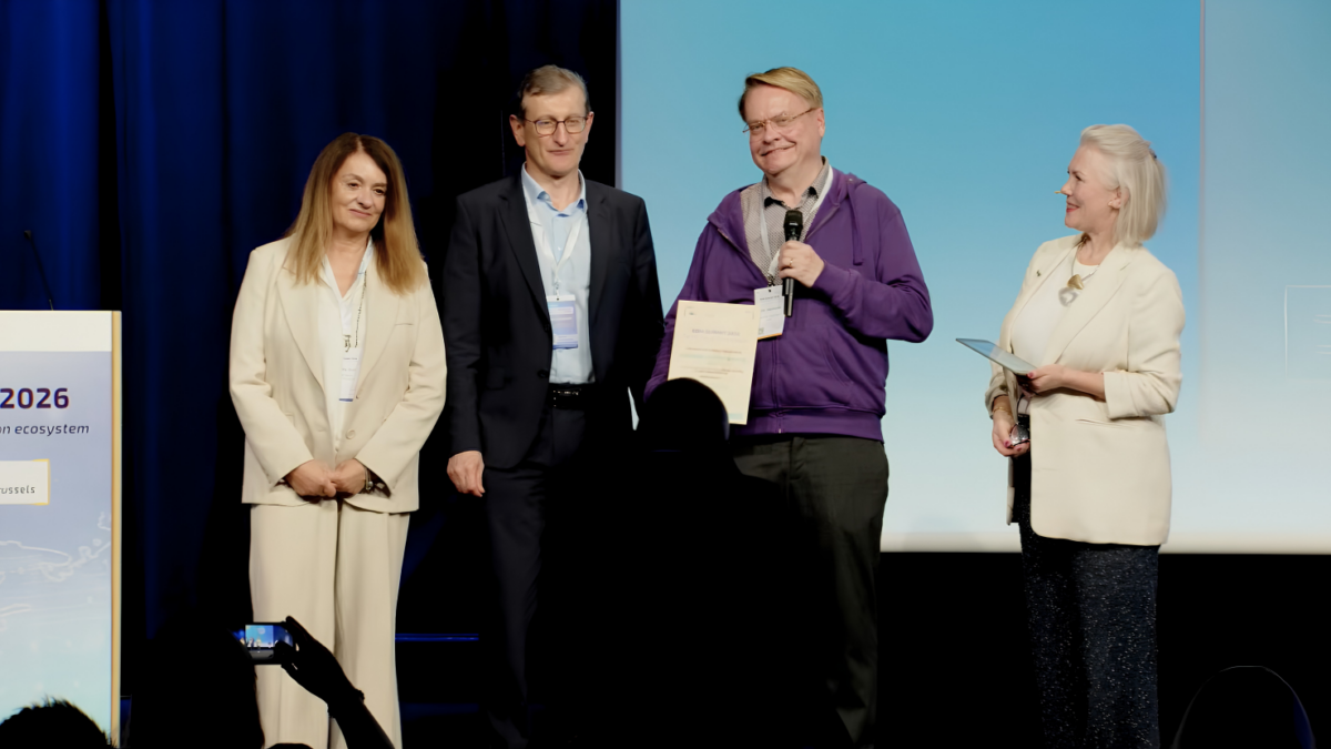 Four people standing on a stage at an awards ceremony; one person holds a certificate and a microphone while the others stand alongside, and another person on the right holds a card or folder. A podium with “2026” is visible on the left, and audience silhouettes appear in the foreground.