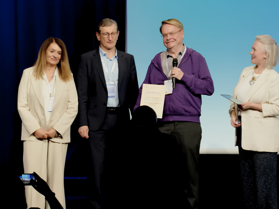 Four people standing on a stage at an awards ceremony; one person holds a certificate and a microphone while the others stand alongside, and another person on the right holds a card or folder. A podium with “2026” is visible on the left, and audience silhouettes appear in the foreground.